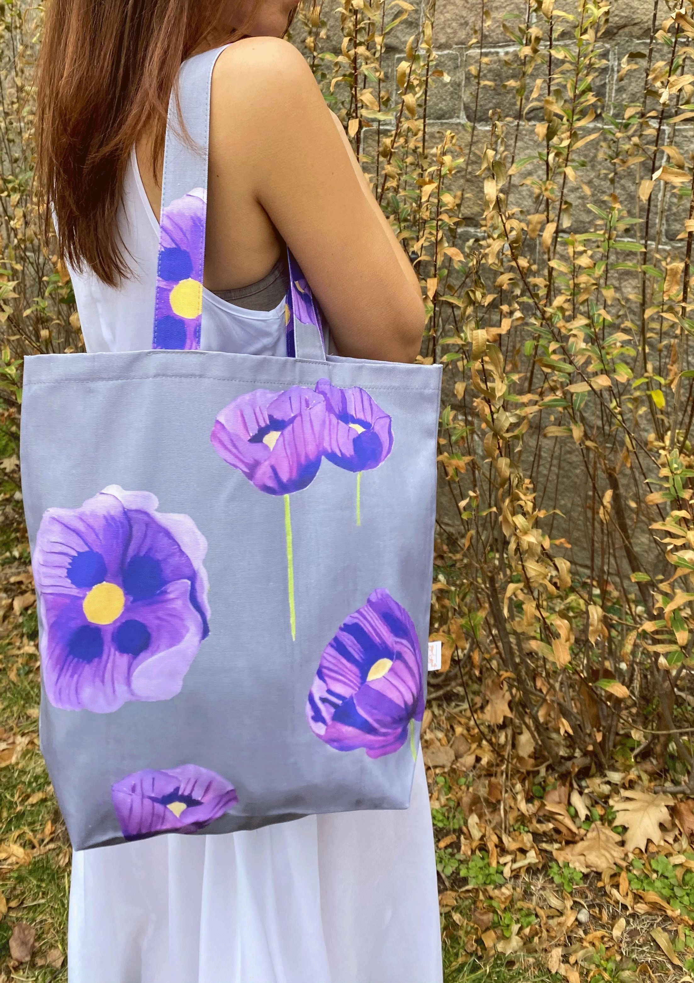 A stylish "Tote Bag: Purple Poppies on Grey," crafted from cotton canvas, hangs on a doorknob, showcasing its beautiful purple poppy design while holding white flowers. Nearby, an umbrella with a wooden handle leans against the door.