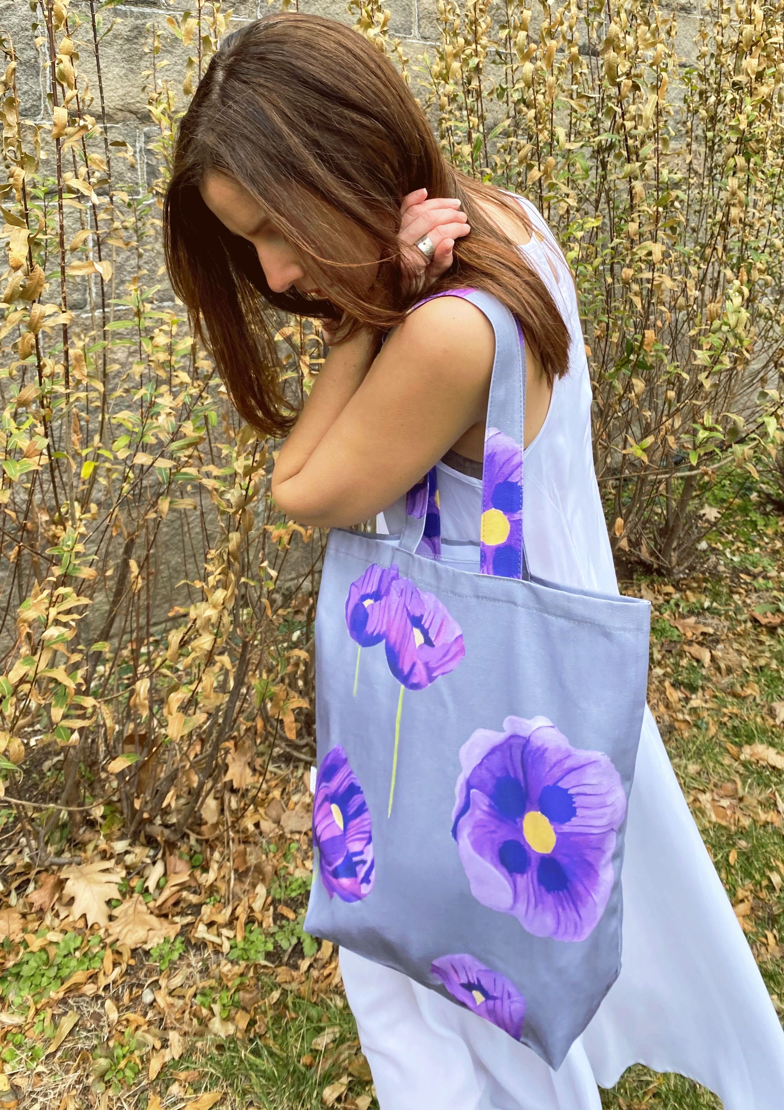 A stylish "Tote Bag: Purple Poppies on Grey," crafted from cotton canvas, hangs on a doorknob, showcasing its beautiful purple poppy design while holding white flowers. Nearby, an umbrella with a wooden handle leans against the door.