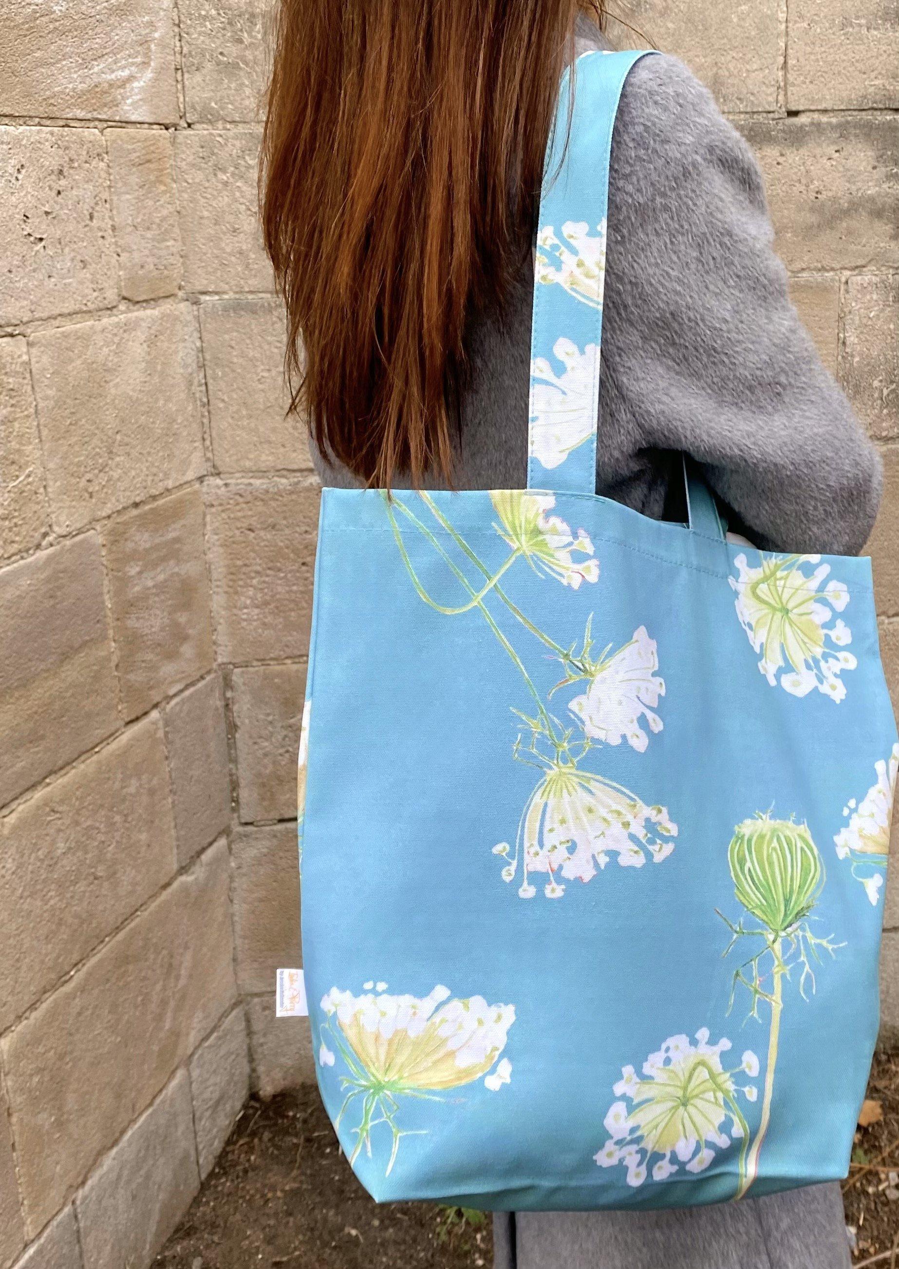 A cotton canvas tote bag featuring a Queen Anne's Lace pattern on a teal background hangs stylishly on a doorknob, next to a folded umbrella. White peonies rest gently on the door, adding an elegant touch to the scene.