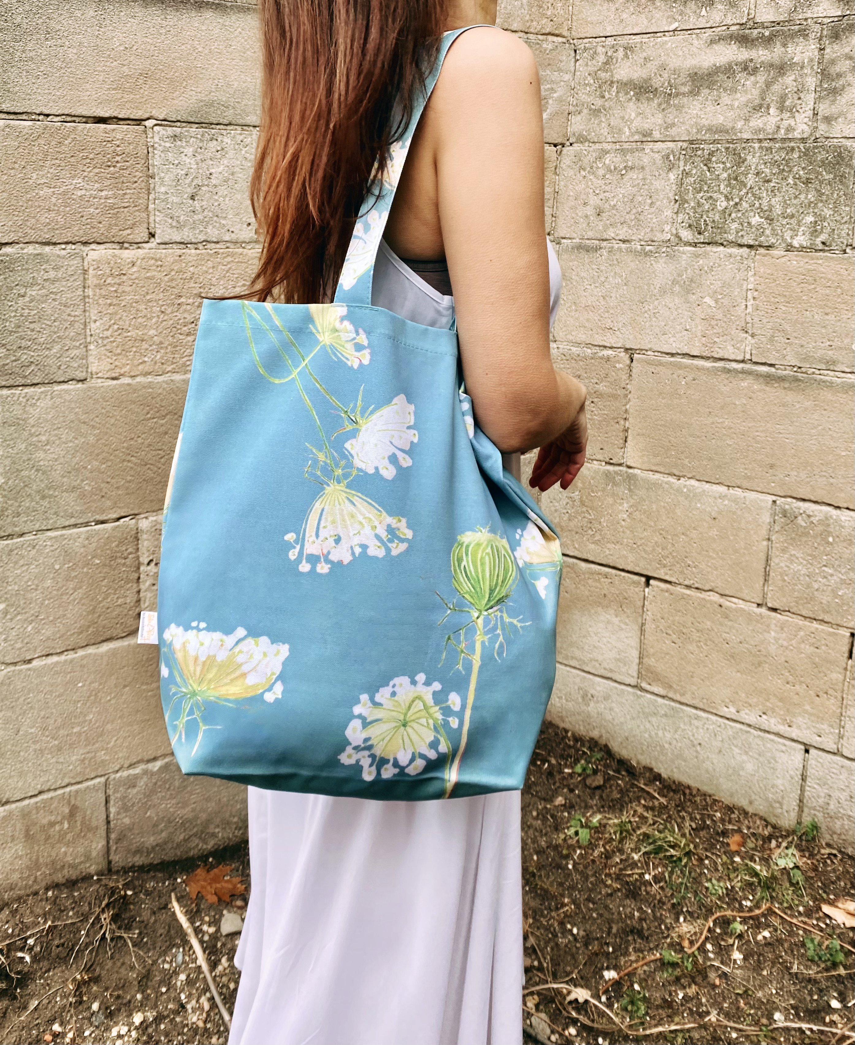 A cotton canvas tote bag featuring a Queen Anne's Lace pattern on a teal background hangs stylishly on a doorknob, next to a folded umbrella. White peonies rest gently on the door, adding an elegant touch to the scene.