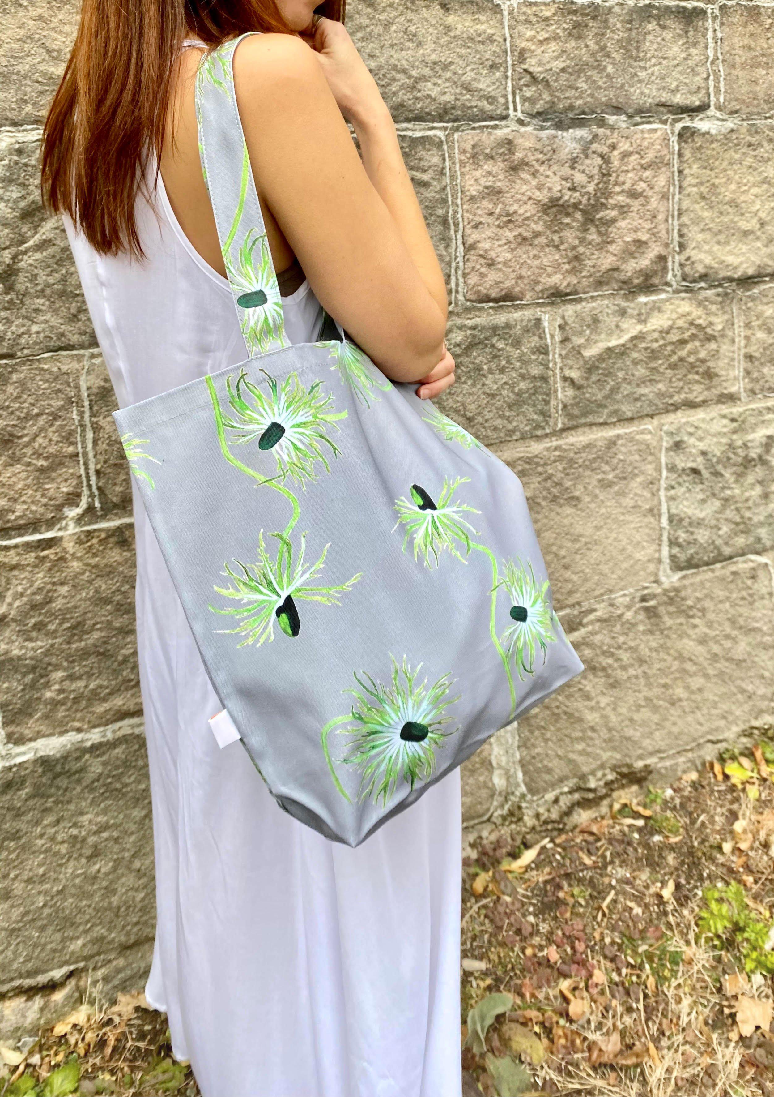 The "Thistle on Grey" tote bag, featuring a gray canvas adorned with floral patterns, hangs elegantly on a doorknob. This stylish shopping tote is filled with pink and white flowers, evoking the charm of an oil-painted scene. Nearby, an umbrella leans against the wall, adding to the picturesque arrangement.