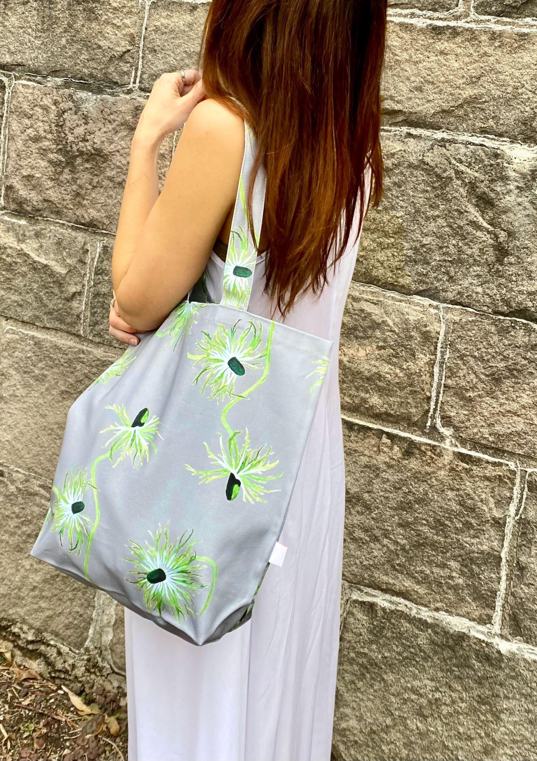 The "Thistle on Grey" tote bag, featuring a gray canvas adorned with floral patterns, hangs elegantly on a doorknob. This stylish shopping tote is filled with pink and white flowers, evoking the charm of an oil-painted scene. Nearby, an umbrella leans against the wall, adding to the picturesque arrangement.