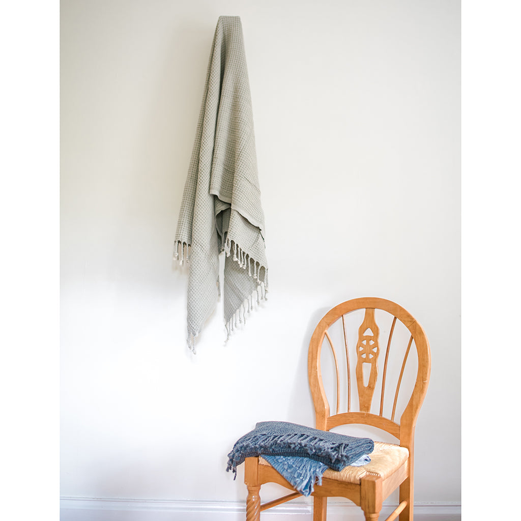 A folded, light gray blanket featuring a waffle texture and fringe tassels at the bottom mirrors the elegance of the Waffle Weave Turkish Towel, combining style and practicality for any cozy setting.