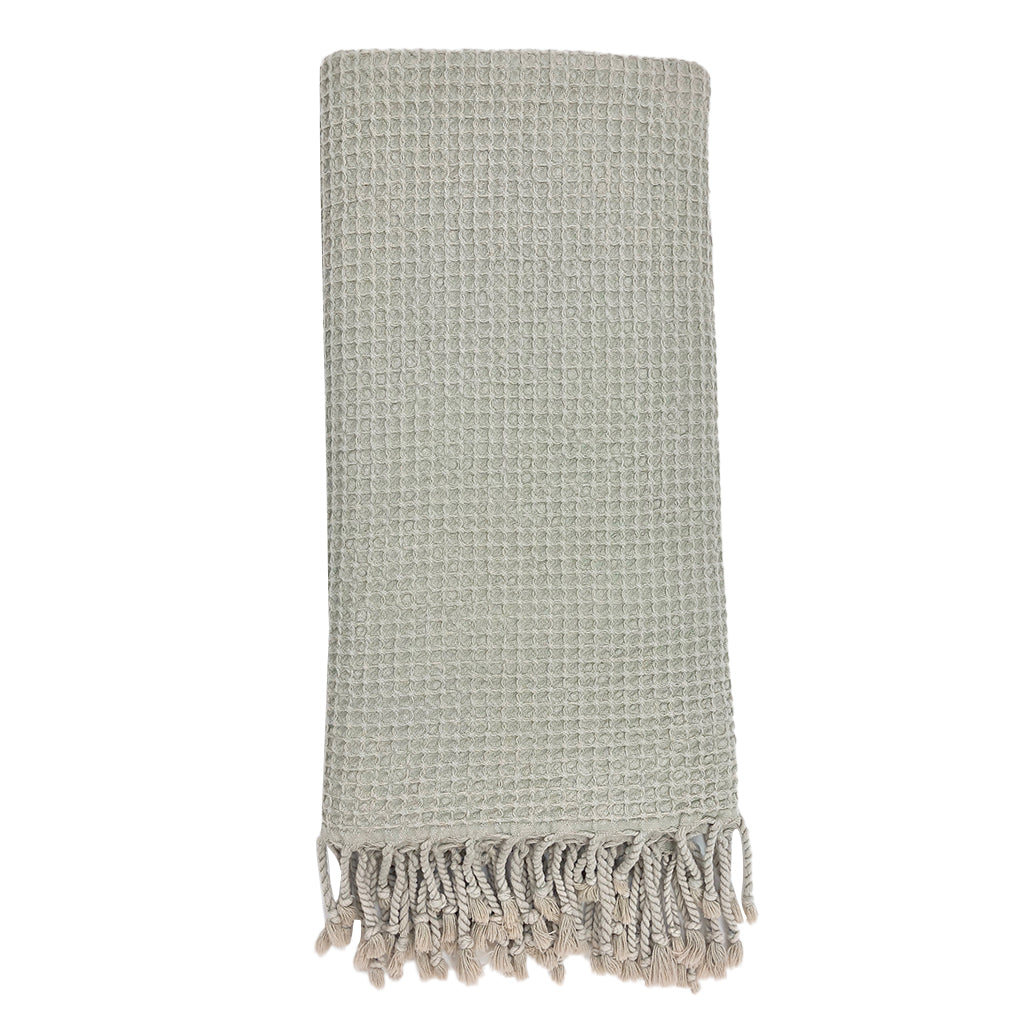 A folded, light gray blanket featuring a waffle texture and fringe tassels at the bottom mirrors the elegance of the Waffle Weave Turkish Towel, combining style and practicality for any cozy setting.