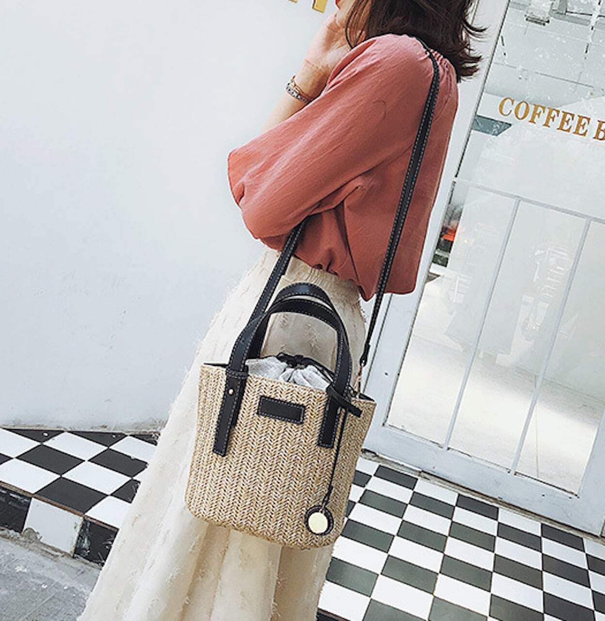 A person wearing a pink blouse and white skirt is carrying a Straw Shoulder Bucket Bag with Vegan Leather Handle, standing near a checkered floor and large windows. This look perfectly embodies summer fashion.