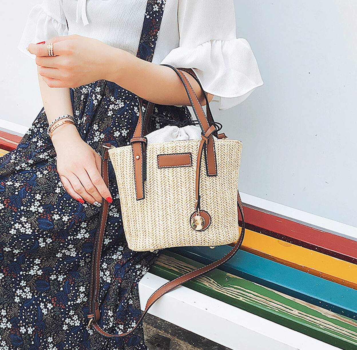 A person wearing a pink blouse and white skirt is carrying a Straw Shoulder Bucket Bag with Vegan Leather Handle, standing near a checkered floor and large windows. This look perfectly embodies summer fashion.