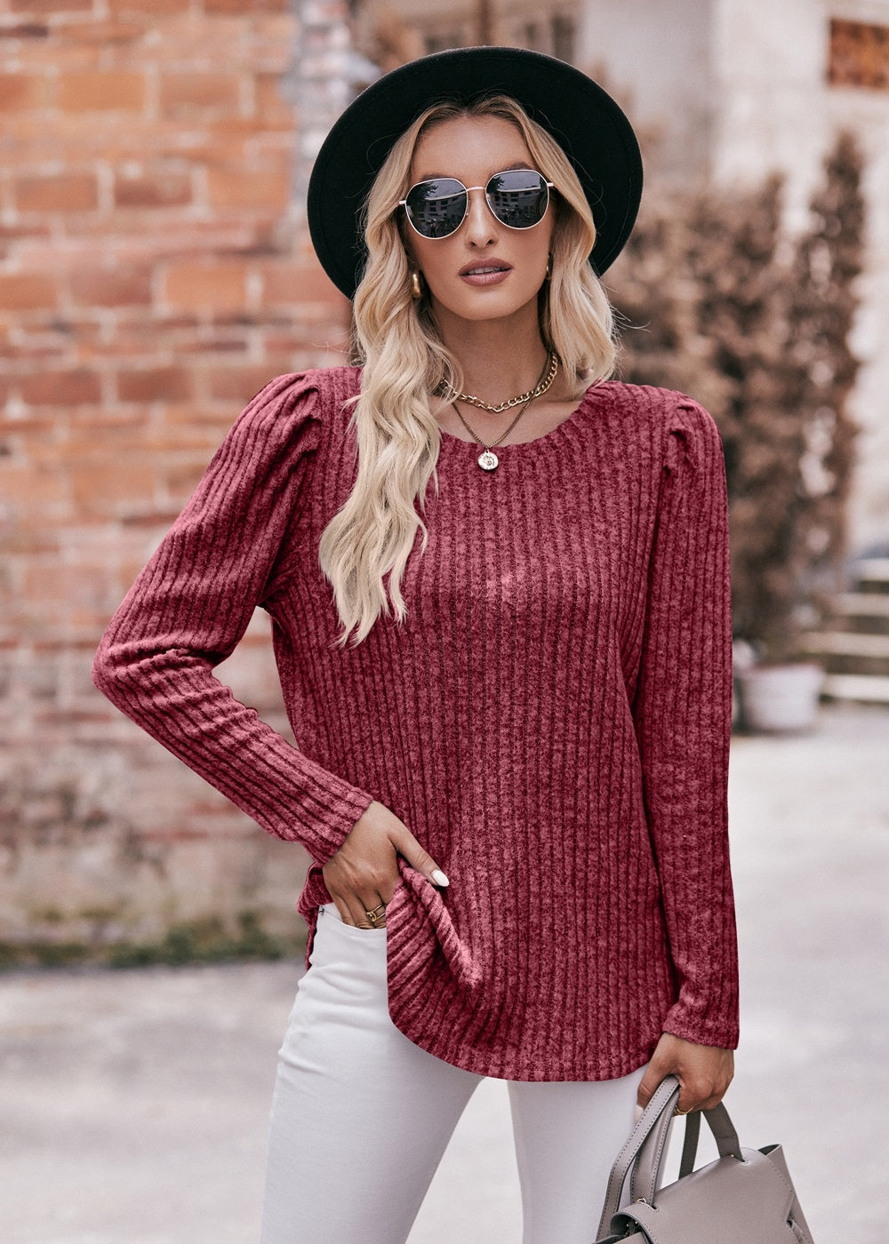 A fashionable woman in a Women's Sweater Puff Long Sleeve Shirt, teal in color, and white pants, accessorized with sunglasses, a necklace, and a black hat, standing in front of a brick wall.