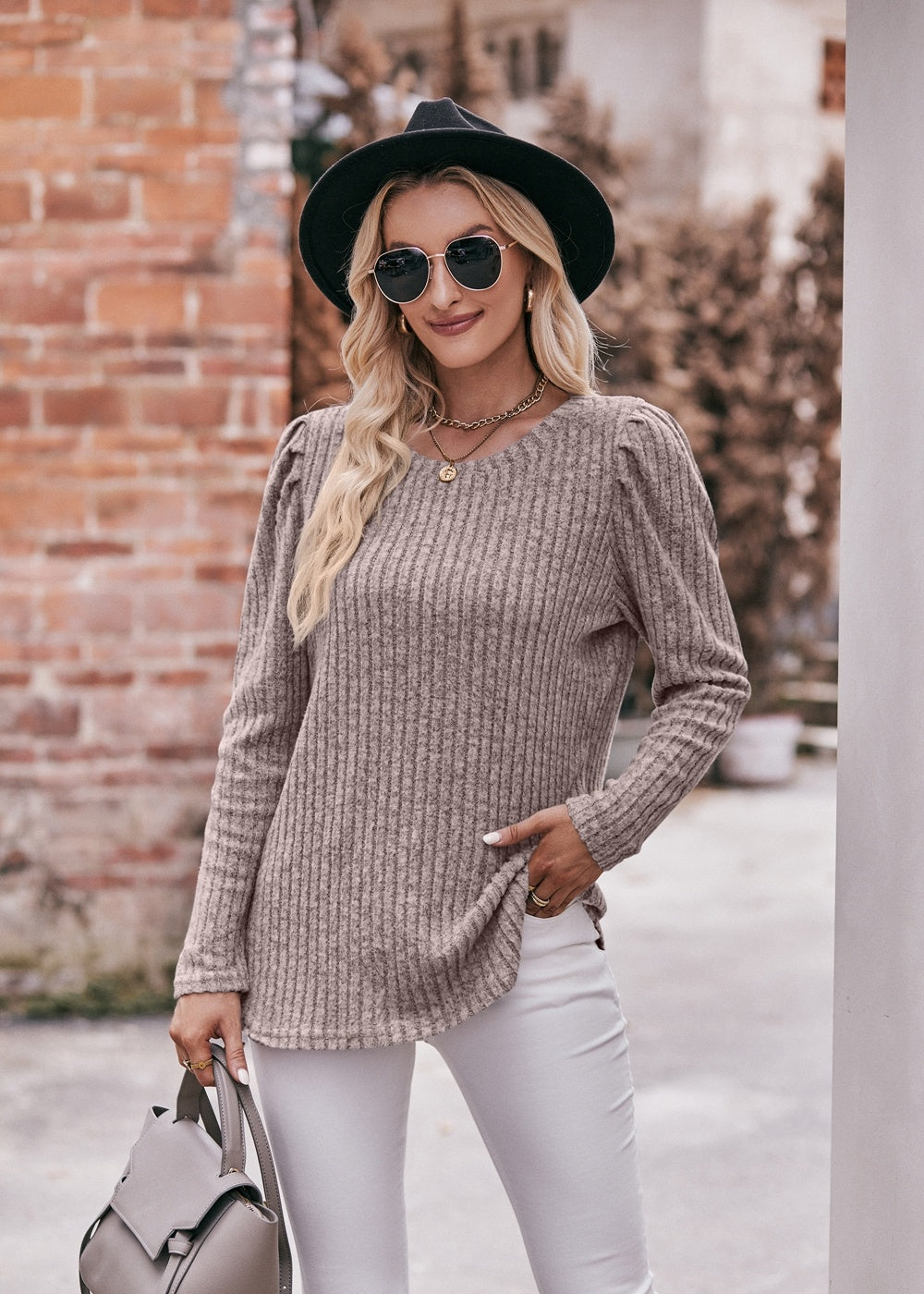 A fashionable woman in a Women's Sweater Puff Long Sleeve Shirt, teal in color, and white pants, accessorized with sunglasses, a necklace, and a black hat, standing in front of a brick wall.