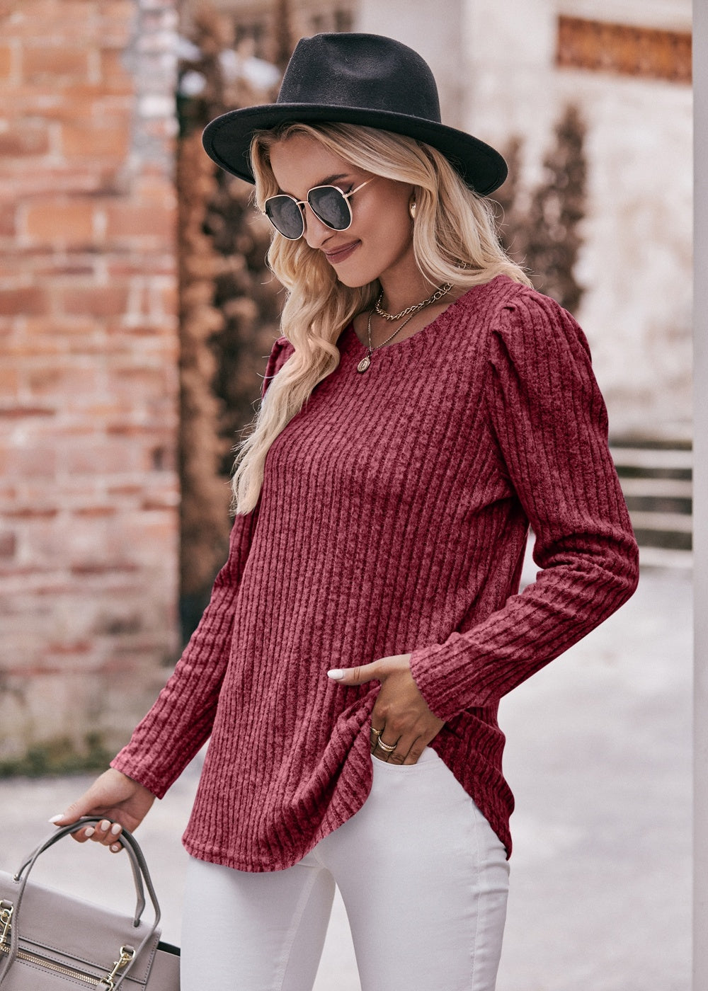 A fashionable woman in a Women's Sweater Puff Long Sleeve Shirt, teal in color, and white pants, accessorized with sunglasses, a necklace, and a black hat, standing in front of a brick wall.