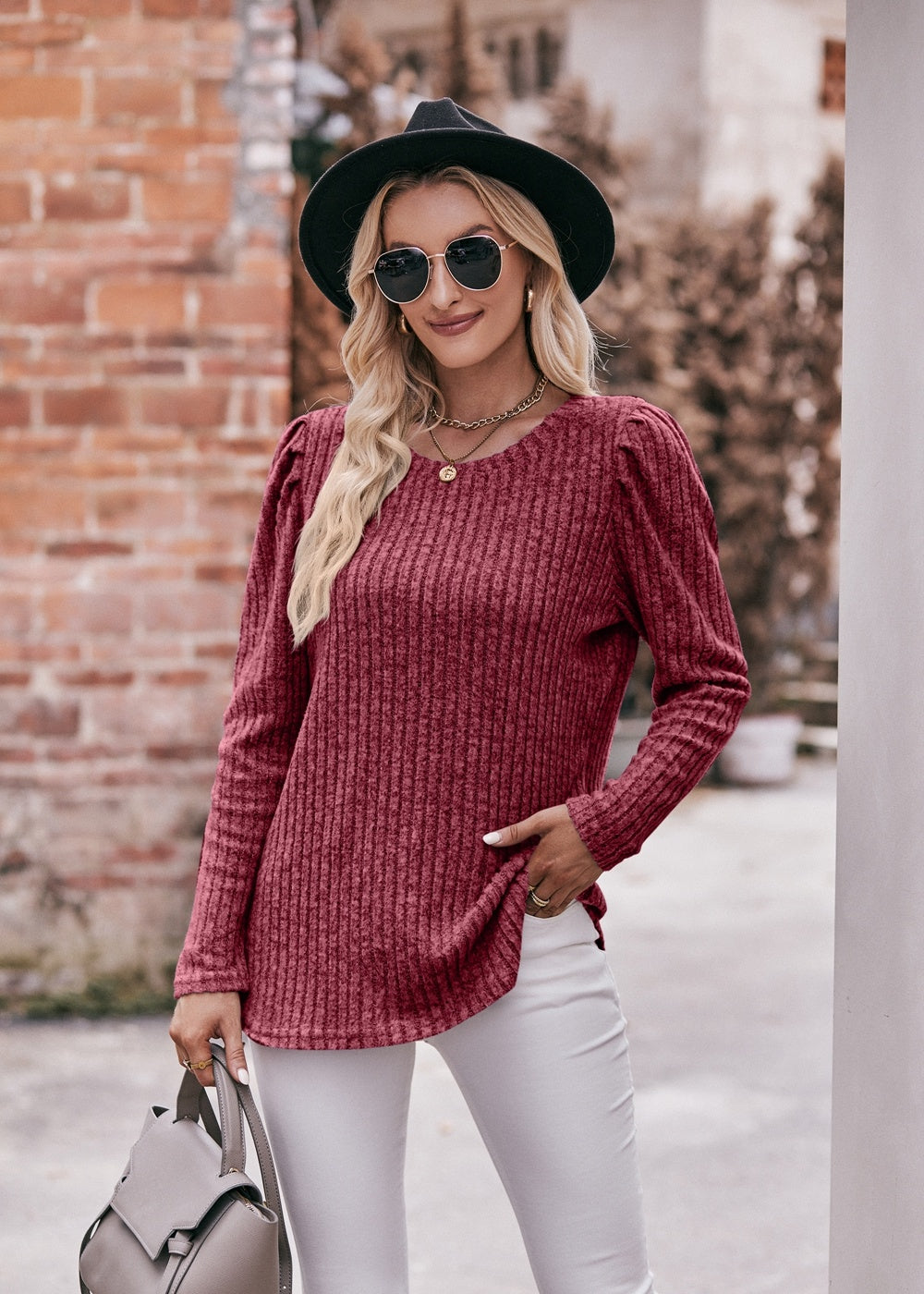 A fashionable woman in a Women's Sweater Puff Long Sleeve Shirt, teal in color, and white pants, accessorized with sunglasses, a necklace, and a black hat, standing in front of a brick wall.