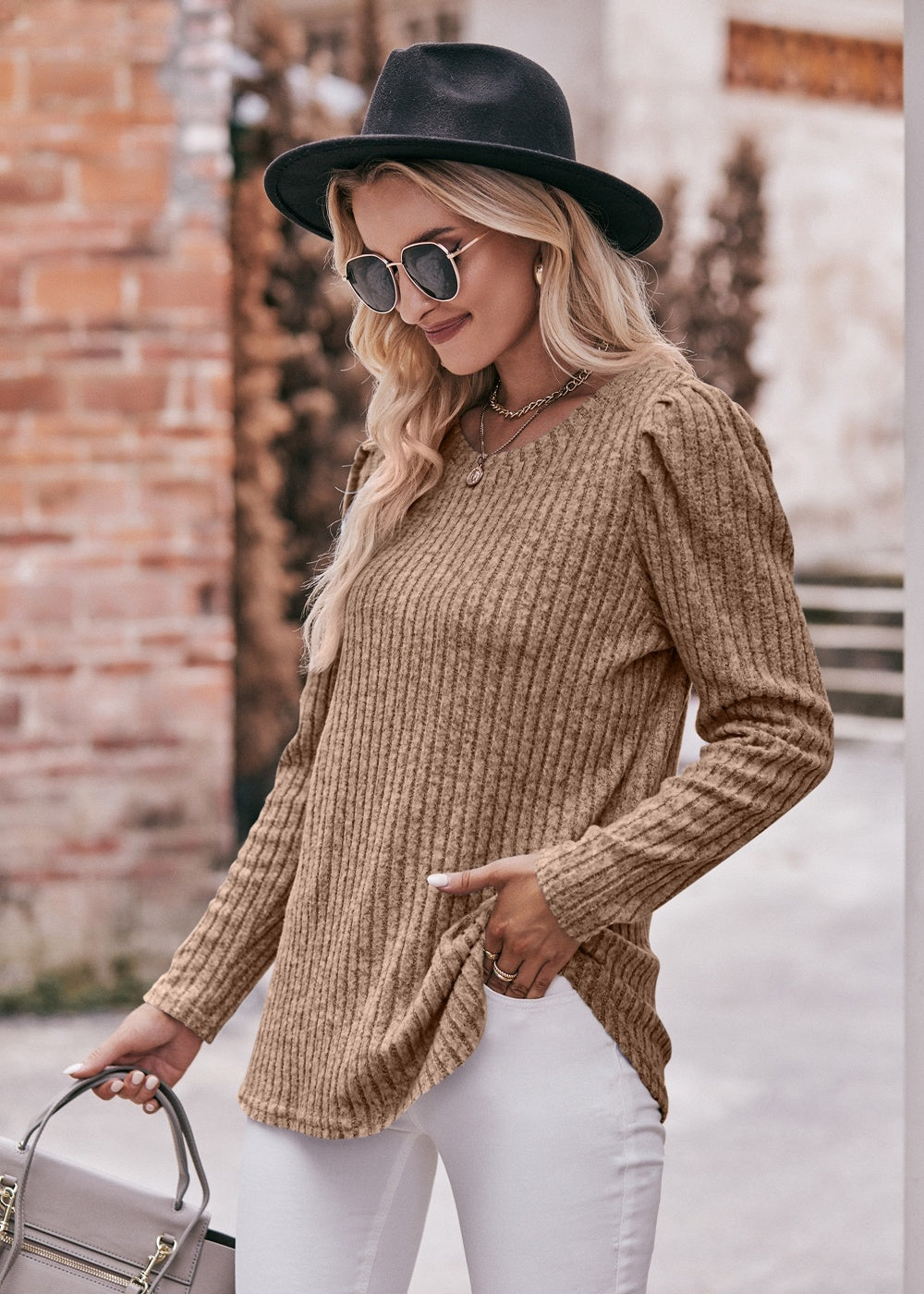 A fashionable woman in a Women's Sweater Puff Long Sleeve Shirt, teal in color, and white pants, accessorized with sunglasses, a necklace, and a black hat, standing in front of a brick wall.