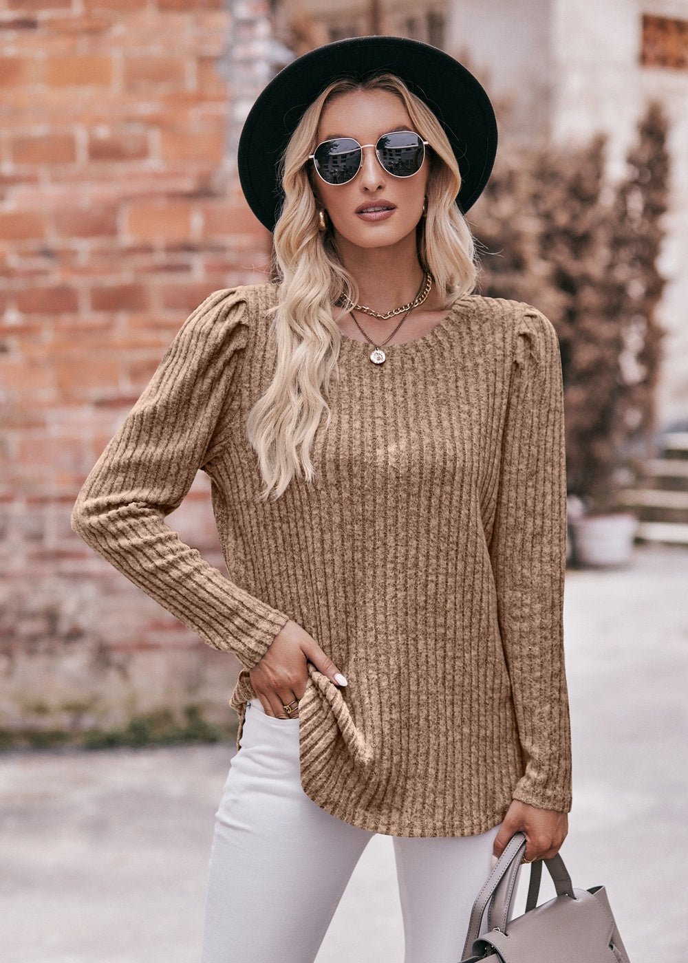 A fashionable woman in a Women's Sweater Puff Long Sleeve Shirt, teal in color, and white pants, accessorized with sunglasses, a necklace, and a black hat, standing in front of a brick wall.