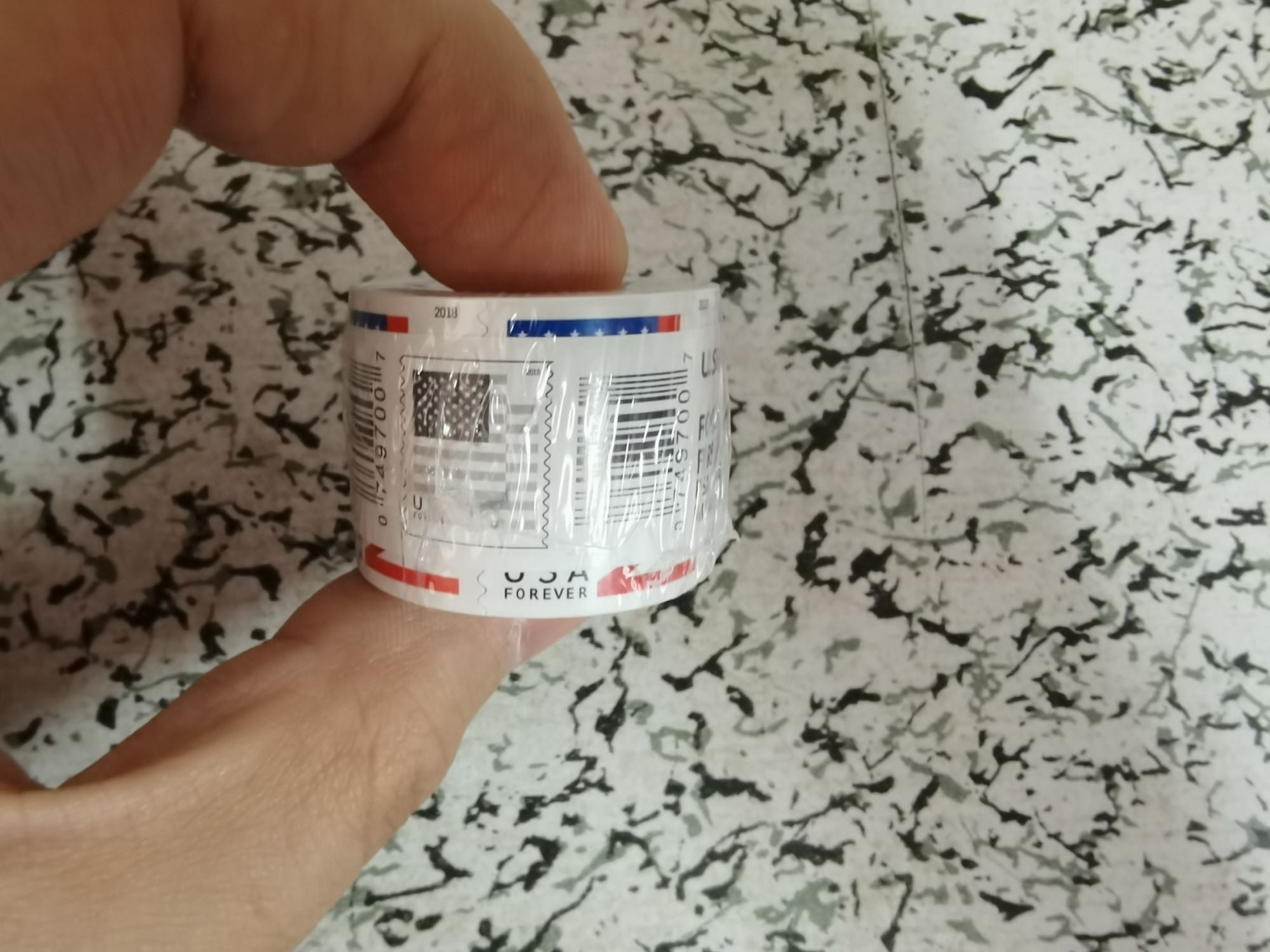 A roll of U.S. Flag 2018 Forever first-class stamps (100 pieces per roll) sits on a brown surface, still wrapped in the original plastic packaging.