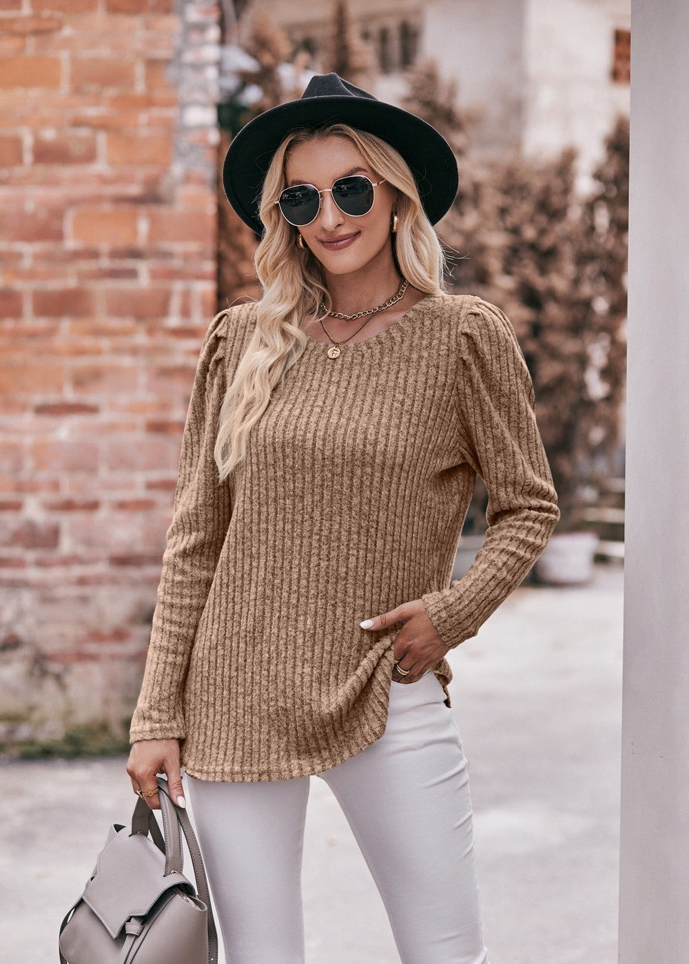 A fashionable woman in a Women's Sweater Puff Long Sleeve Shirt, teal in color, and white pants, accessorized with sunglasses, a necklace, and a black hat, standing in front of a brick wall.