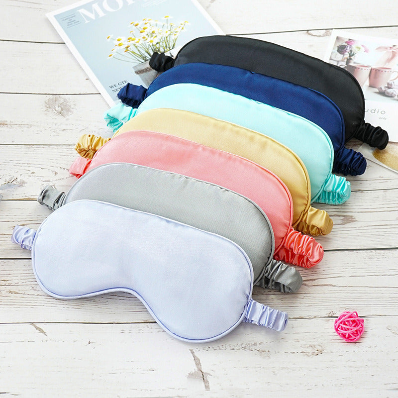 A collection of colorful Imitated Silk Eye Patch Shading Sleep Eye Masks and hair scrunchies arranged on a wooden surface next to magazines and decorative flowers.