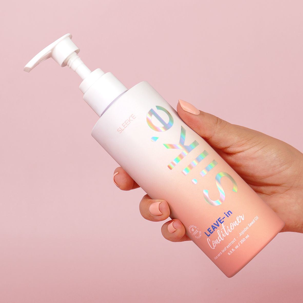 A sleek'd hand holding a bottle of Silk'e leave In Conditioner on a pink background.