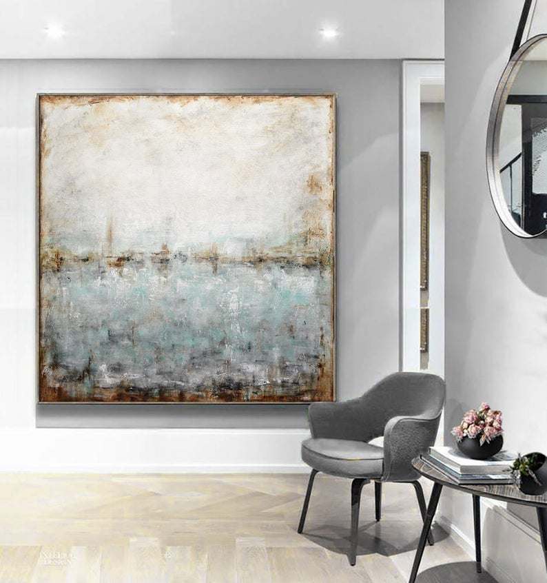 Abstract painting in muted tones on a gallery wall, with a modern grey chair and small table with flowers to the right, in a minimalist interior.