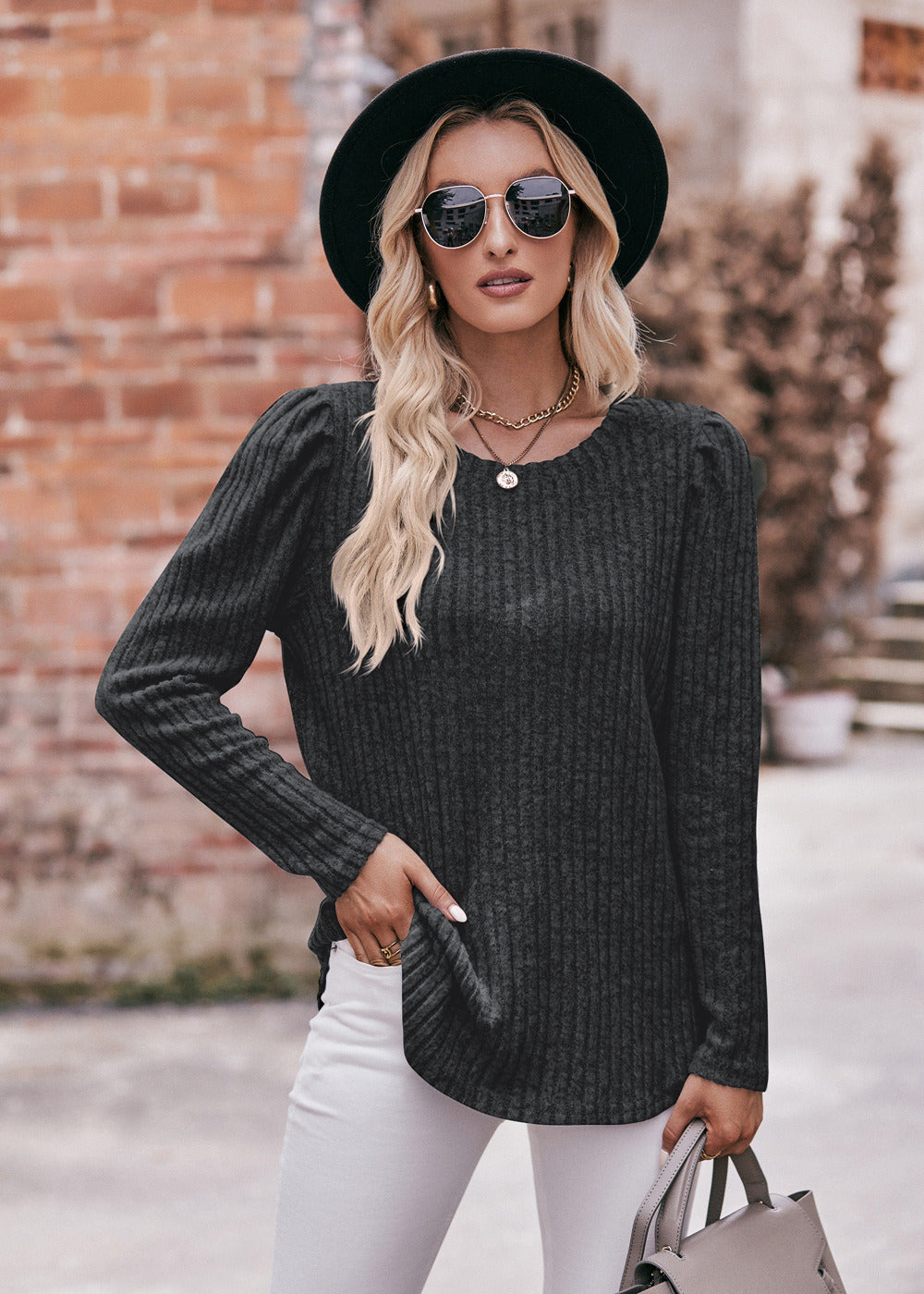 A fashionable woman in a Women's Sweater Puff Long Sleeve Shirt, teal in color, and white pants, accessorized with sunglasses, a necklace, and a black hat, standing in front of a brick wall.