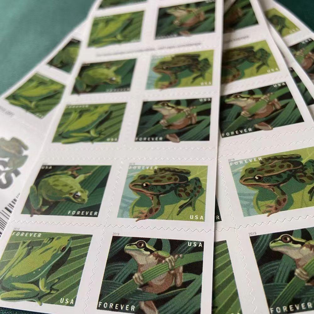 A stack of Frogs 2019 - 5 Booklets / 100 Pcs postage stamps featuring various green frogs illustrations scattered on a white surface, showcasing details of the frogs dating back to 2019.