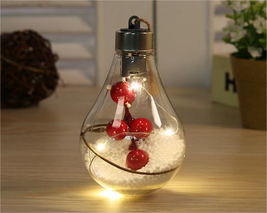 Five LED-transparent Christmas decoration balls from the "Pack Of 5 Led Transparent Christmas Decoration Ball; Creative Simulation Light Bulb Christmas Tree Decoration Pendant Plastic Ball" pack, filled with festive elements like berries and pine branches, are displayed on top of a closed book in front of a white shuttered background.