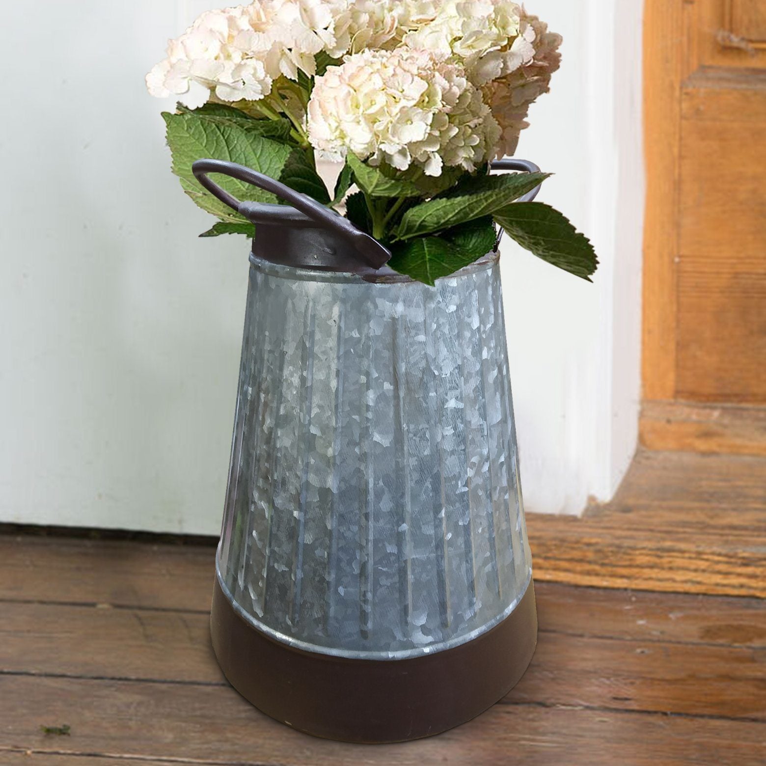 A vintage Galvanized Metal Corrugated Flower Vase with Curved Side Handles, filled with hydrangea flowers, positioned against a wooden door and white wall.