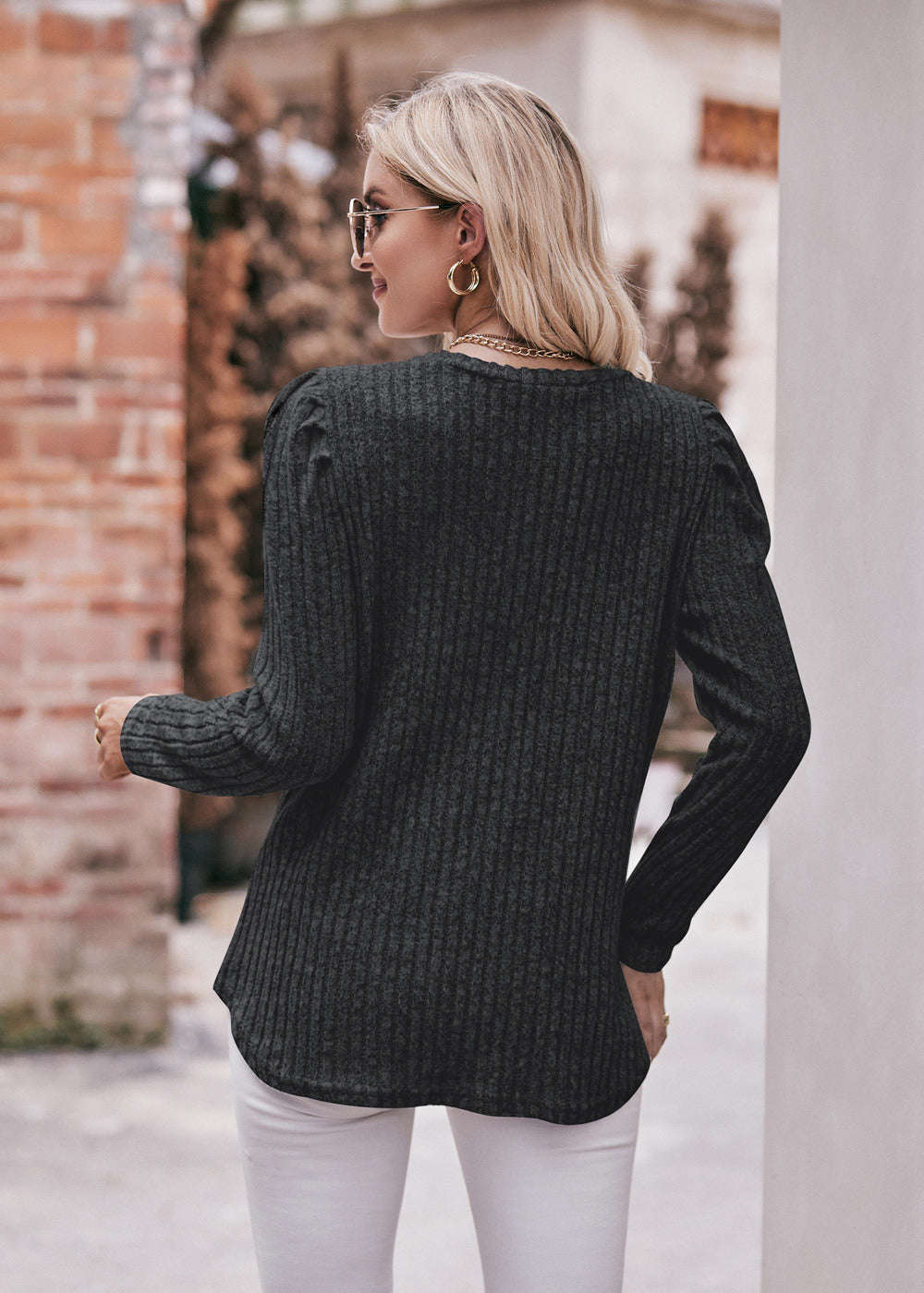 A fashionable woman in a Women's Sweater Puff Long Sleeve Shirt, teal in color, and white pants, accessorized with sunglasses, a necklace, and a black hat, standing in front of a brick wall.