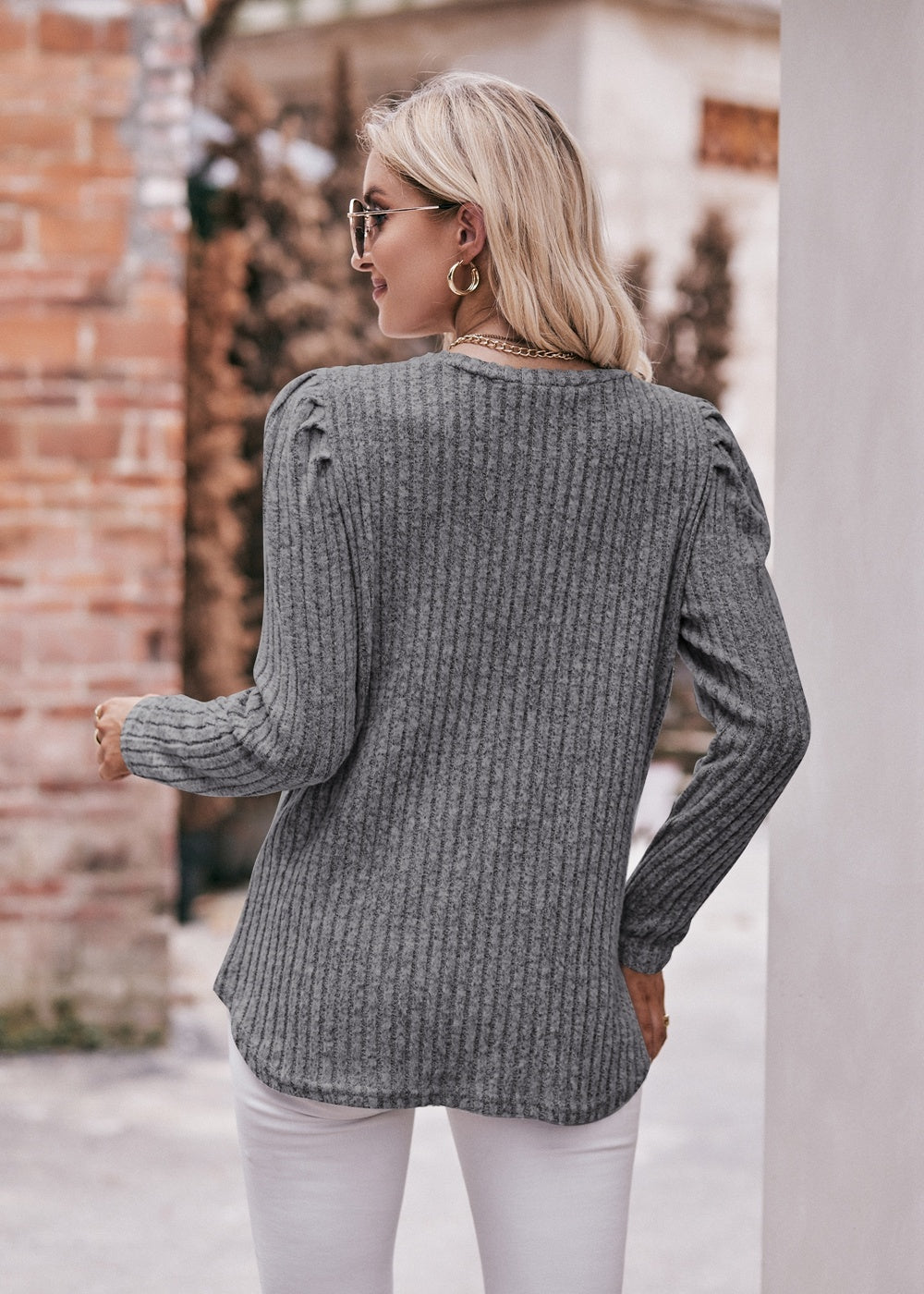 A fashionable woman in a Women's Sweater Puff Long Sleeve Shirt, teal in color, and white pants, accessorized with sunglasses, a necklace, and a black hat, standing in front of a brick wall.