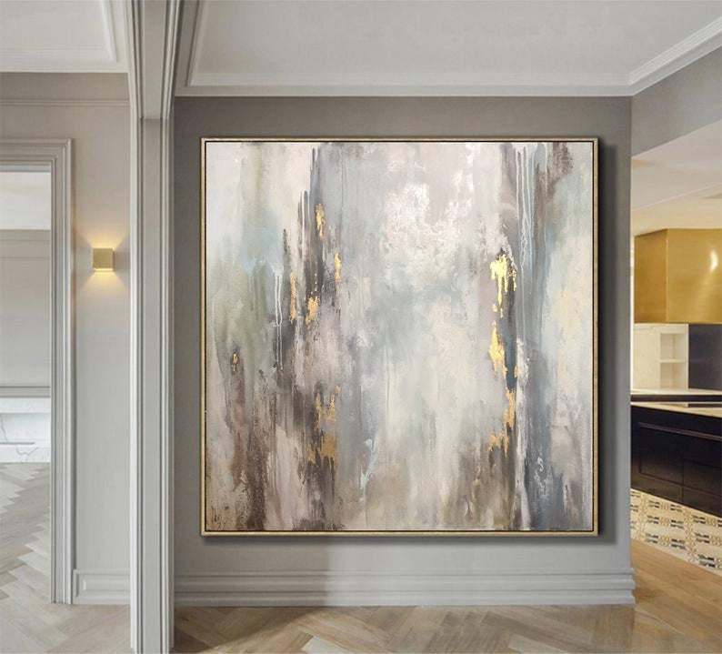 A large Abstract Paintings On Canvas with gray, gold, and white brush strokes, framed and hanging on a wall in a modern room with elegant decor.