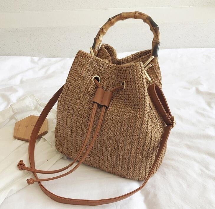 A drawstring straw backpack with a bamboo handle, a leather strap, and a drawstring closure, placed on a white surface.
