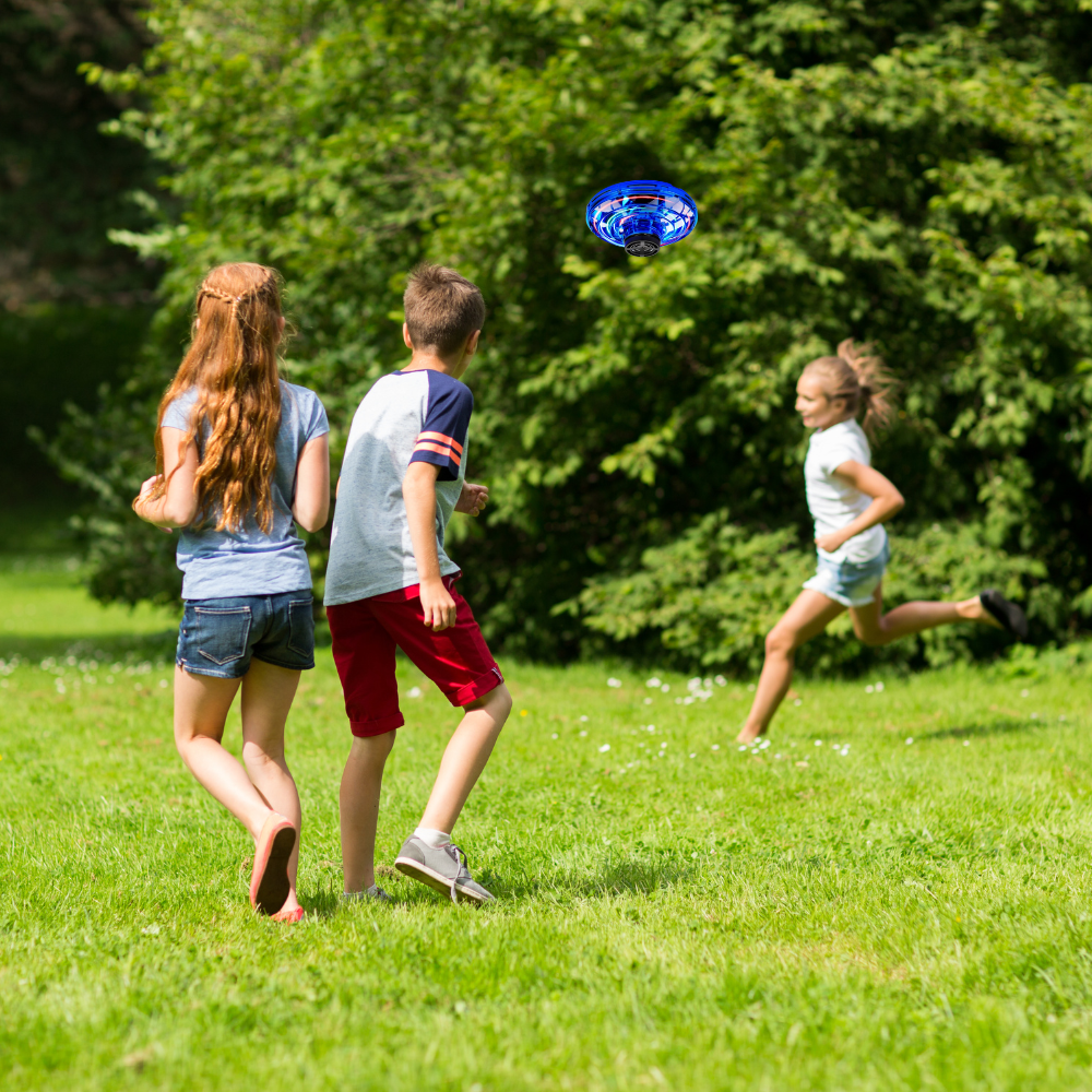 Children playing tag in a lush green park, with one girl in blue shorts reaching to tag a boy in a striped shirt, while a Glow Drone 360 Flying Toy with LED lights hovers above.