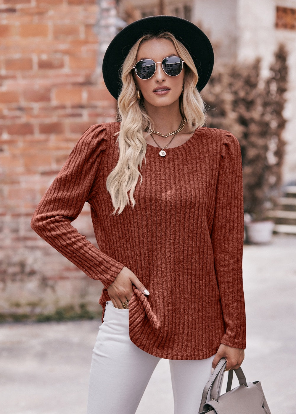 A fashionable woman in a Women's Sweater Puff Long Sleeve Shirt, teal in color, and white pants, accessorized with sunglasses, a necklace, and a black hat, standing in front of a brick wall.