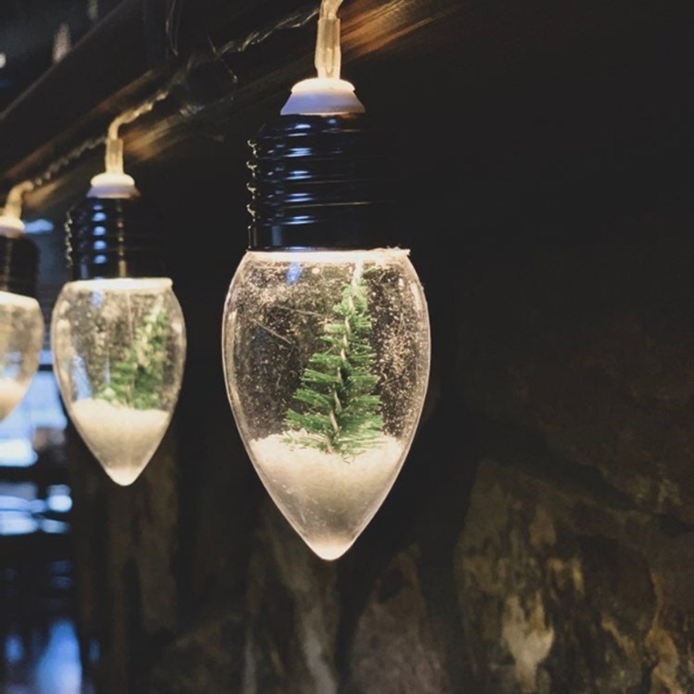 Miniature Christmas Trees encased in clear, teardrop-shaped light bulbs, illuminated by battery-powered lights and hanging from a dark ceiling.