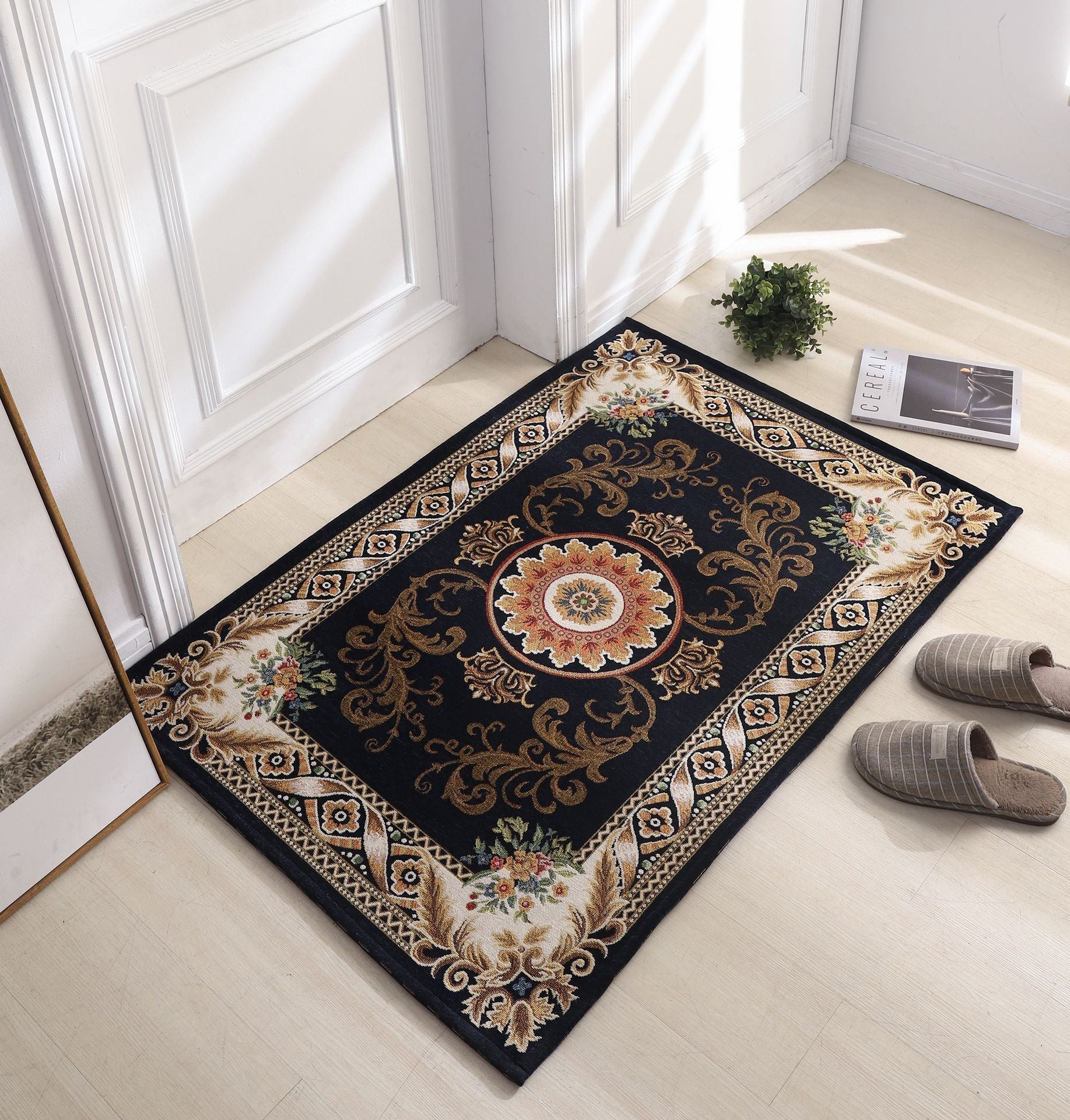 A small, ornate Dornier woven carpet with an intricate floral design in red and brown hues lies on a light-colored wooden floor next to gray slippers. A small plant and a book are placed nearby against a white wall.