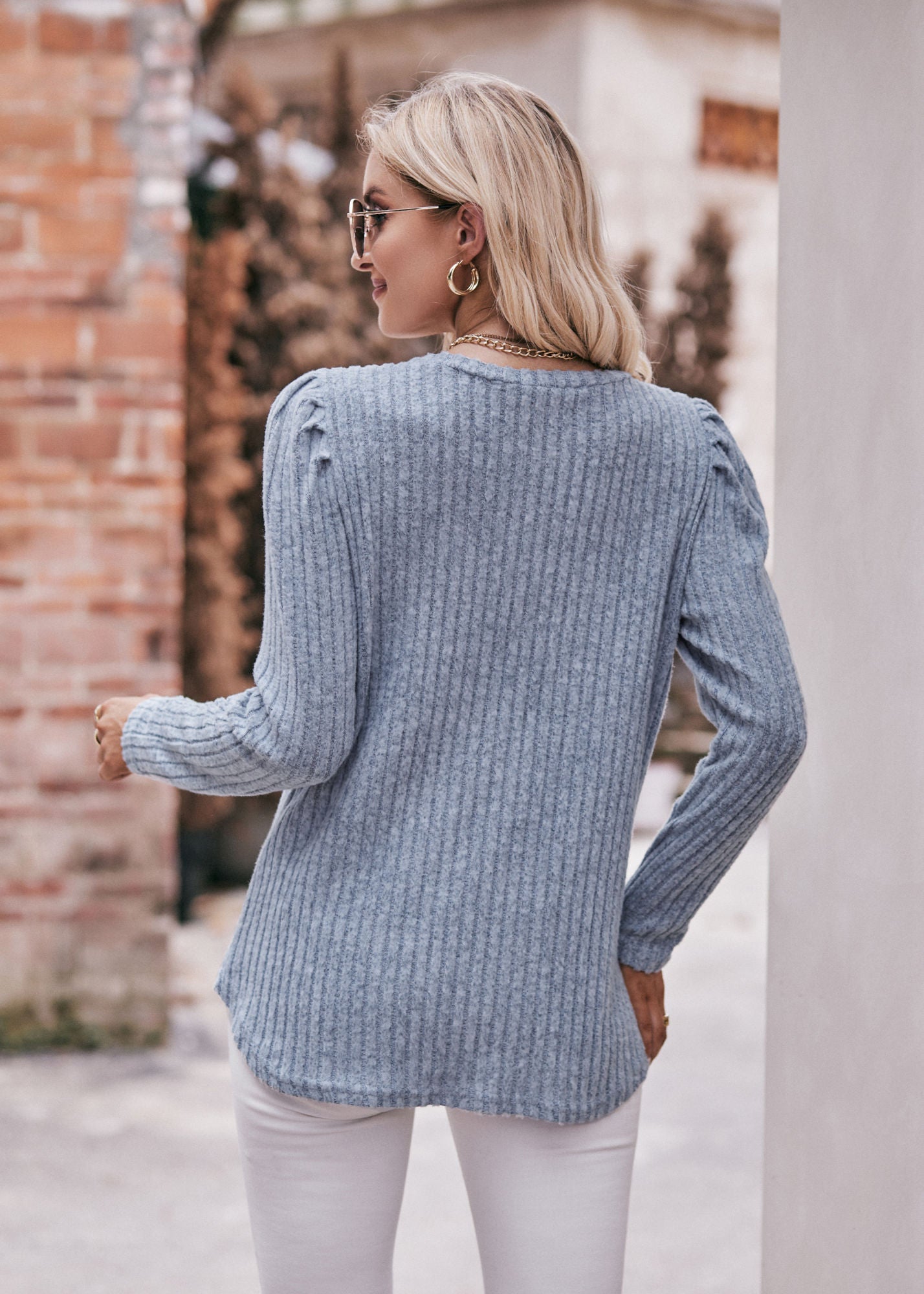 A fashionable woman in a Women's Sweater Puff Long Sleeve Shirt, teal in color, and white pants, accessorized with sunglasses, a necklace, and a black hat, standing in front of a brick wall.